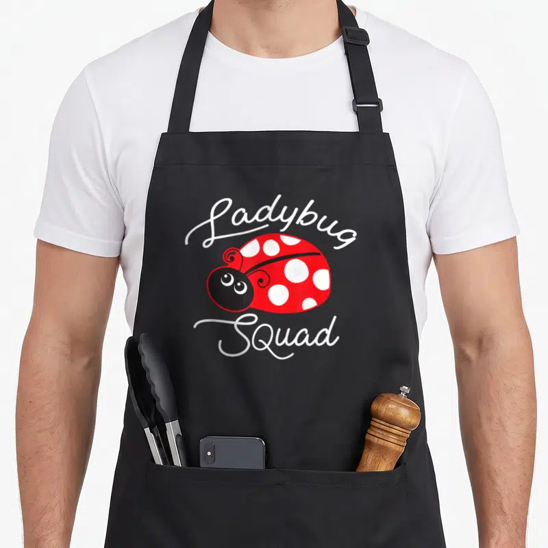 Funny squad ladybug apron - Cartoon red ladybug with white spots centered between white script reading Ladybug Squad — shown on a black cotton canvas apron against a white background