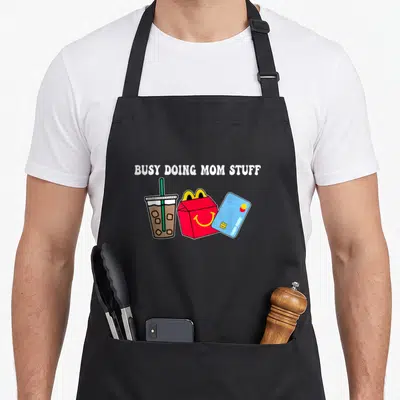 Busy mother's day aprons - BUSY DOING MOM STUFF text above iced drink, red fry box, and blue credit card cartoon — shown on a black cotton canvas apron against a white background