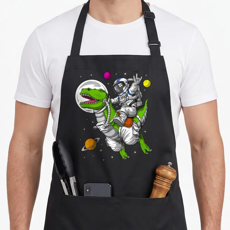 Space dinosaur apron - Cartoon astronaut riding green t-rex in space with pink yellow and orange planets — shown on a black cotton canvas apron against a white background