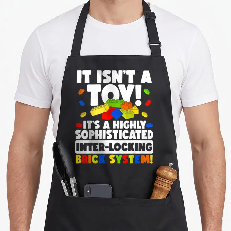 Builder brick grill master apron - Scattered red yellow blue green orange plastic bricks with sarcastic inter-locking — shown on a black cotton canvas apron against a white background