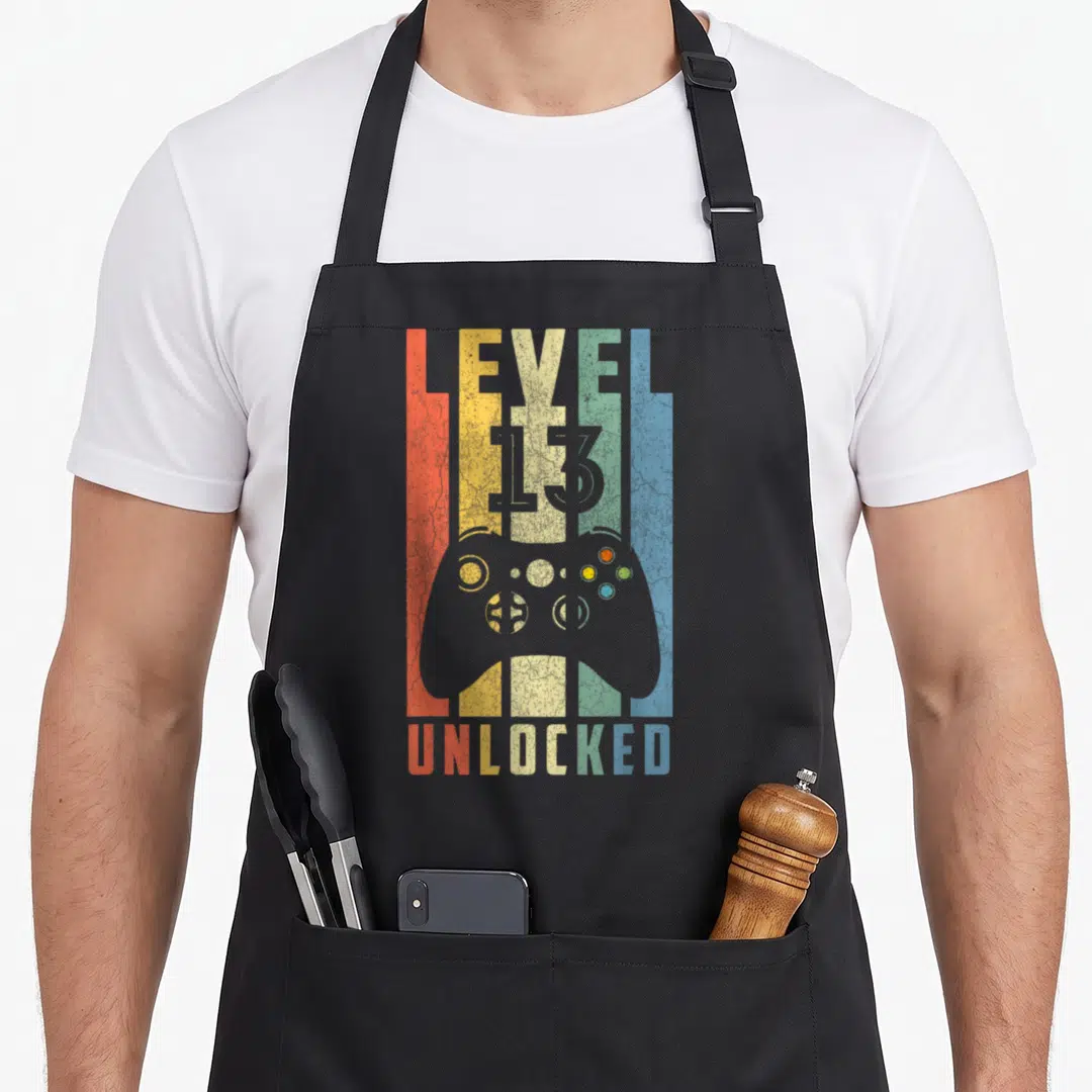 Unlocked 13th birthday apron - Retro game controller silhouette over five colored stripes with LEVEL 13 UNLOCKED text — shown on a black cotton canvas apron against a white background