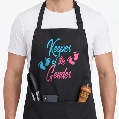 Keeper baby gender reveal apron - Stacked Keeper of the Gender text with two blue and two pink footprints — shown on a black cotton canvas apron against a white background
