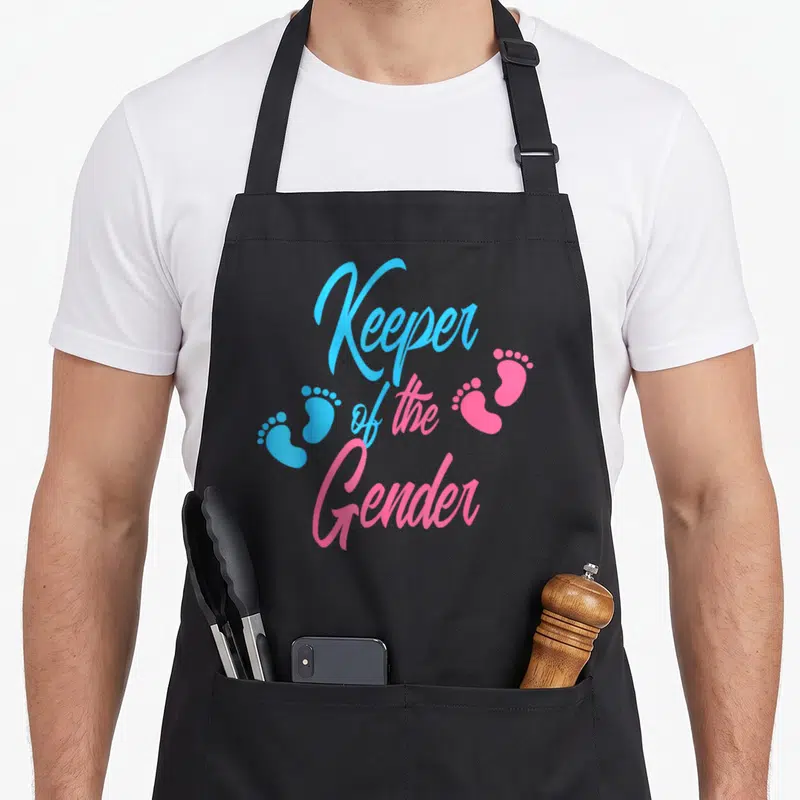Keeper baby gender reveal apron - Stacked Keeper of the Gender text with two blue and two pink footprints — shown on a black cotton canvas apron against a white background