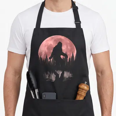 Night bigfoot apron - Grey Bigfoot silhouette walking in front of a pink moon and forest with water reflection — shown on a black cotton canvas apron against a white background