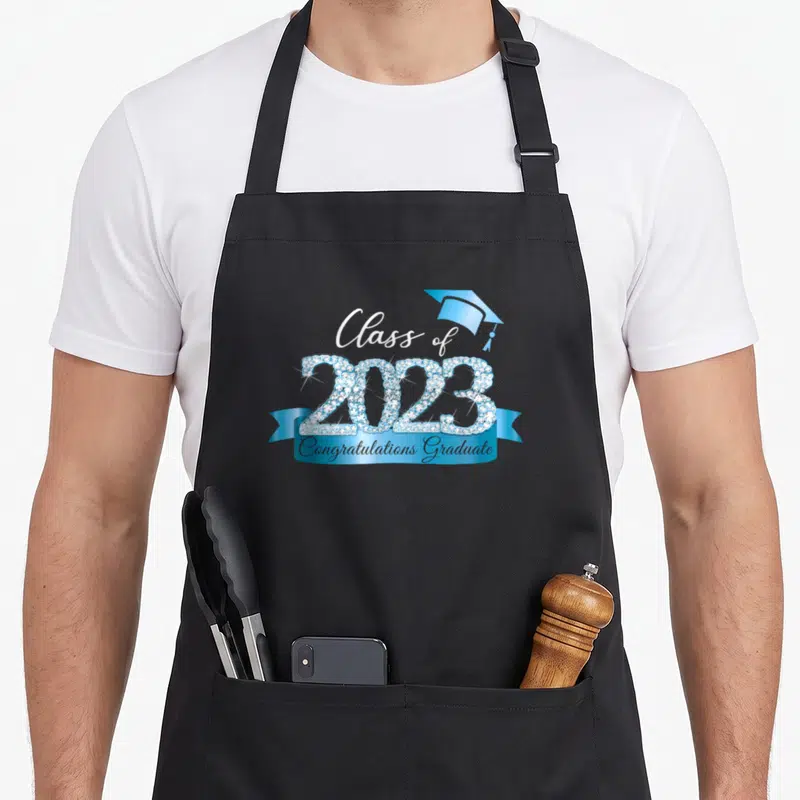 Class congratulations apron - Blue rhinestone 2023 text with ribbon and cap celebrating graduate — shown on a black cotton canvas apron against a white background