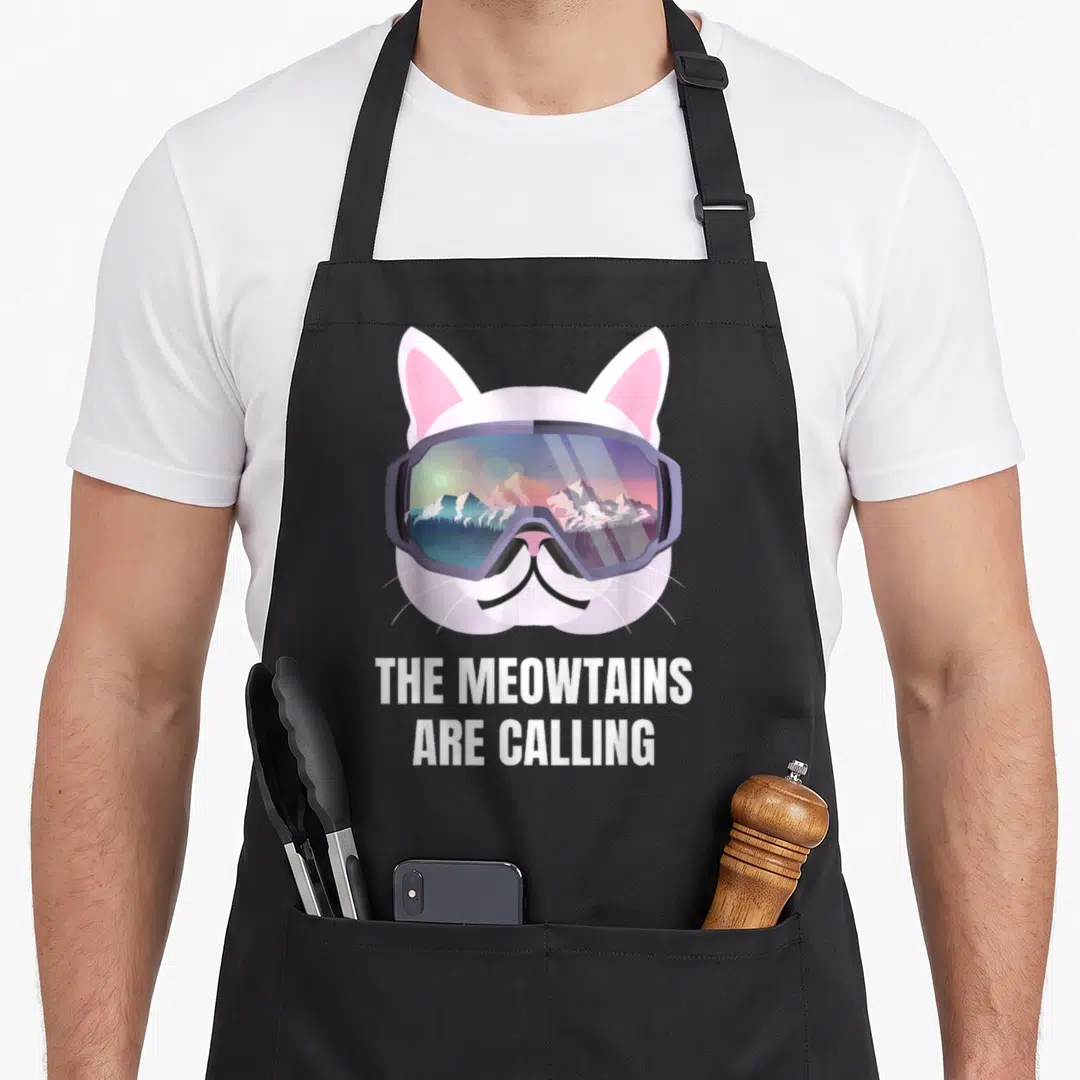 Ski goggles and apron - Cartoon cat head in purple goggles reflecting mountains above white text THE MEOWTAINS ARE — shown on a black cotton canvas apron against a white background