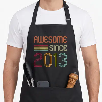 Funny 11th birthday apron - Retro text AWESOME SINCE 2013 in orange red yellow teal beige stacked vertically — shown on a black cotton canvas apron against a white background