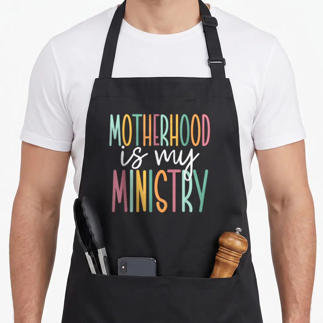 Ministry motherhood apron - Stacked text MOTHERHOOD is my MINISTRY in mint yellow pink orange and white — shown on a black cotton canvas apron against a white background