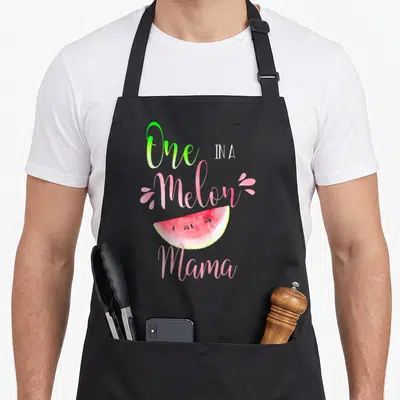 Funny melon birthday apron - One IN A Melon Mama watermelon slice watercolor text pink green — shown on a black cotton canvas apron against a white background