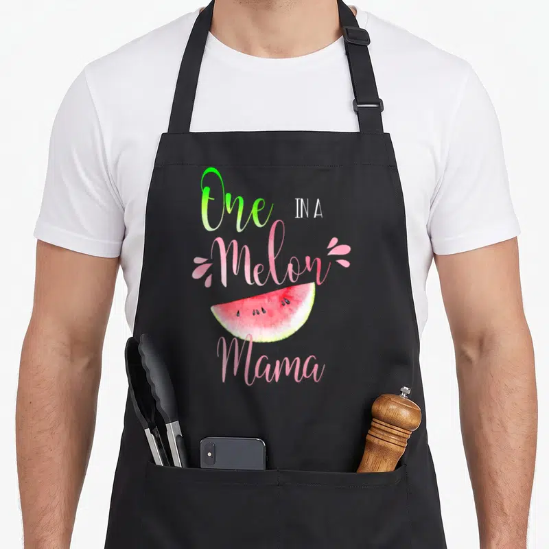 Funny melon birthday apron - One IN A Melon Mama watermelon slice watercolor text pink green — shown on a black cotton canvas apron against a white background