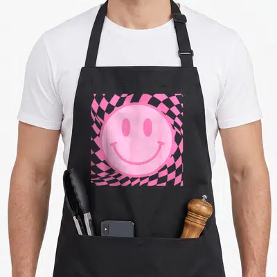 Smile smiling face on apron - Large pink smiley face centered on distorted pink and grey checkerboard pattern — shown on a black cotton canvas apron against a white background
