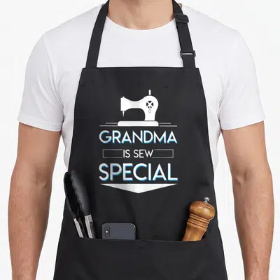 Sew grandma apron - White sewing machine above stacked text Grandma Is Sew Special with blue outlines — shown on a black cotton canvas apron against a white background