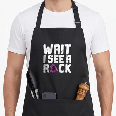 Rock geologist apron - White distressed text WAIT I SEE A ROCK with purple agate slice replacing the O — shown on a black cotton canvas apron against a white background
