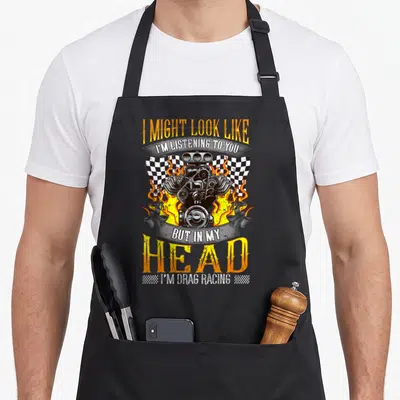 Fan racing apron - Retro engine with flames and checkered flags under sarcastic drag racing text — shown on a black cotton canvas apron against a white background