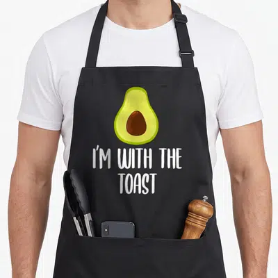 Halloween couple toast apron - Cartoon avocado with brown pit above white I'M WITH THE TOAST text — shown on a black cotton canvas apron against a white background