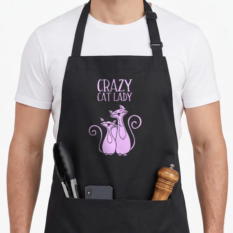 Vintage crazy cat lady apron - Two purple cartoon cats standing side-by-side below centered text reading CRAZY CAT LADY — shown on a black cotton canvas apron against a white background