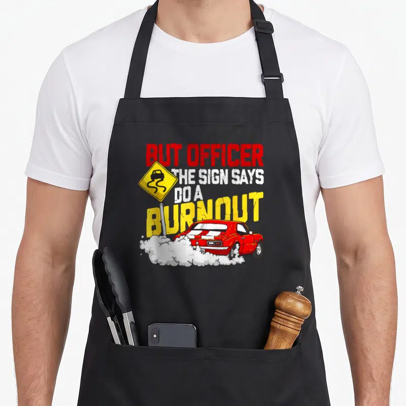 Burnout car apron - Red muscle car doing burnout with white smoke, yellow sign, and sarcastic text — shown on a black cotton canvas apron against a white background
