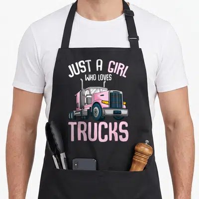 Vintage trucker semi truck apron - Pink semi-truck cab illustration with arched text JUST A GIRL WHO LOVES TRUCKS — shown on a black cotton canvas apron against a white background