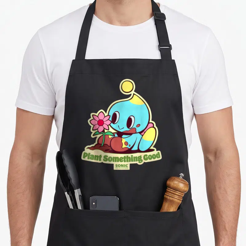 Earth plant apron - Blue Chao holding pink flower in brown dirt with Plant Something Good text — shown on a black cotton canvas apron against a white background