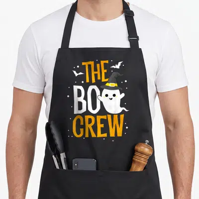 Boo crew halloween apron - Cartoon ghost wearing a witch hat with THE BOO CREW text and scattered dots — shown on a black cotton canvas apron against a white background