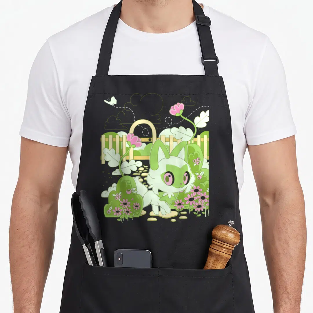 Sprigatito nature apron - Cartoon cat-like creature in a garden with pink flowers, fence, and flying butterfly in green — shown on a black cotton canvas apron against a white background