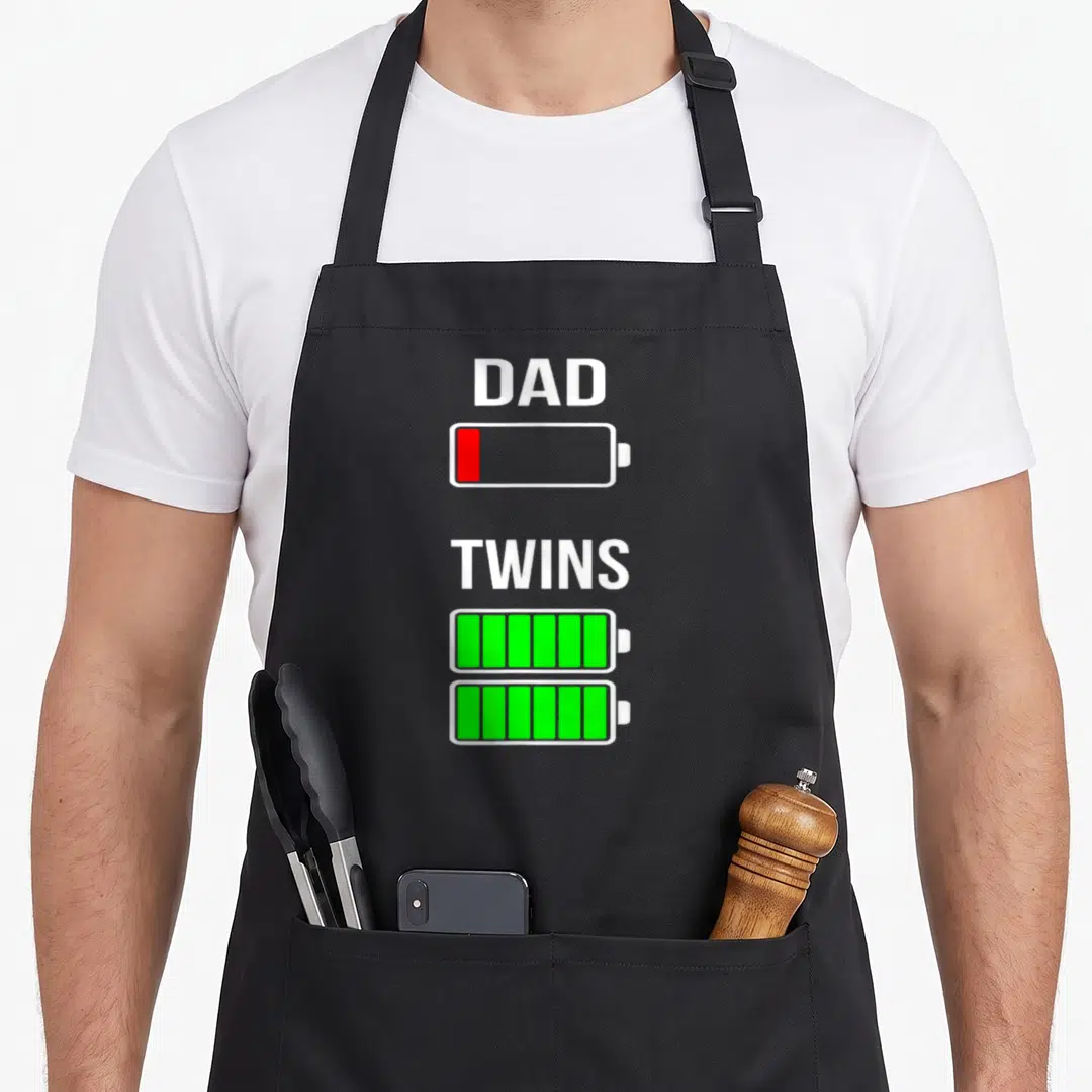 Tired battery apron - DAD above red low battery, TWINS above two full green batteries — shown on a black cotton canvas apron against a white background