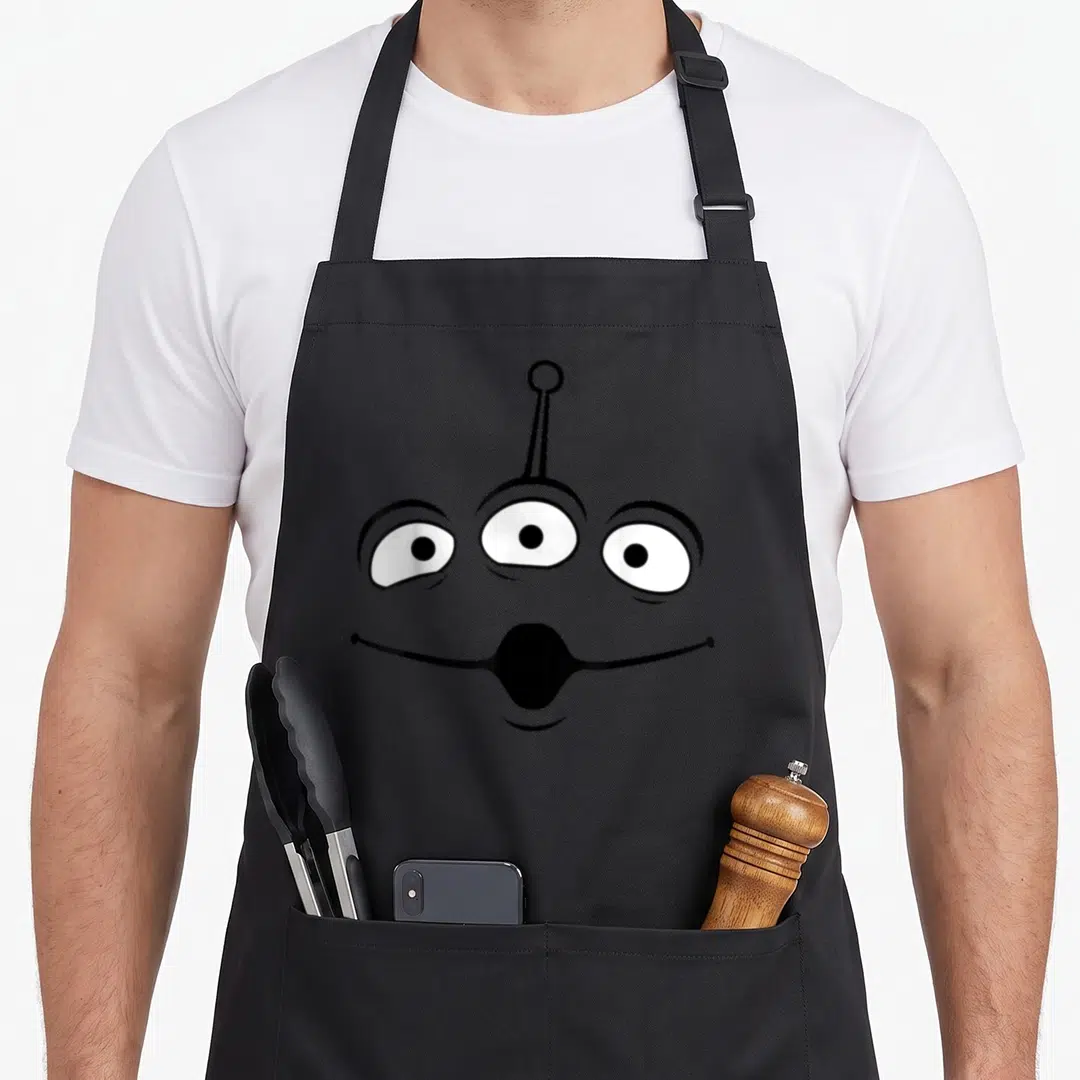 Alien halloween apron - Grey cartoon alien face with three eyes and antenna on black background — shown on a black cotton canvas apron against a white background