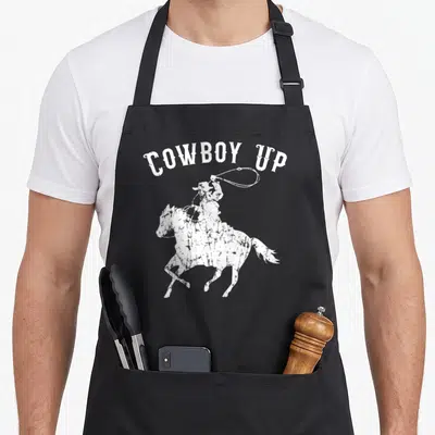 Fan western aprons - Distressed white cowboy silhouette on horseback swinging lasso with arched COWBOY UP text — shown on a black cotton canvas apron against a white background