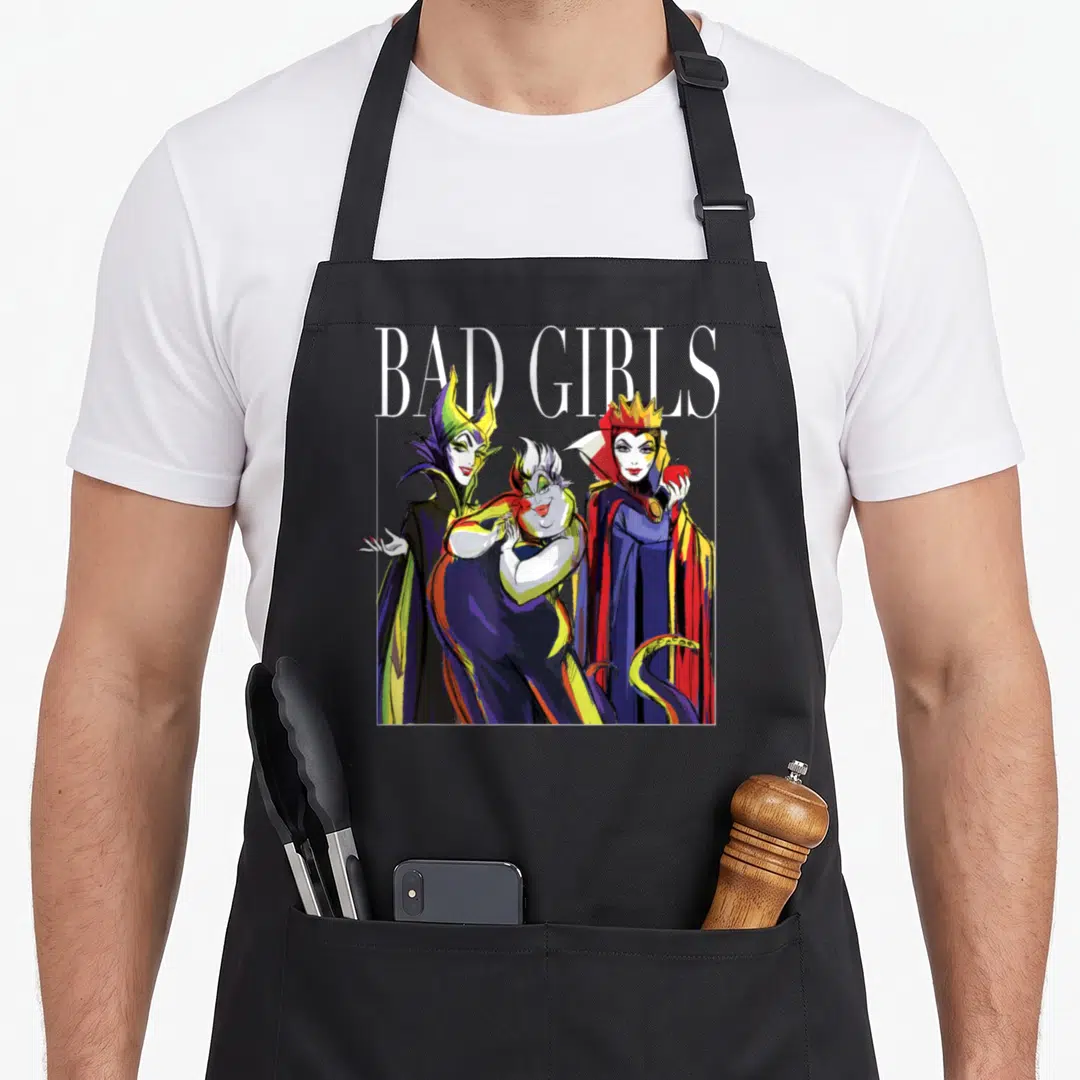 Villain the painted apron - BAD GIRLS text above three overlapping cartoon female villains in purple blue red and yellow — shown on a black cotton canvas apron against a white background