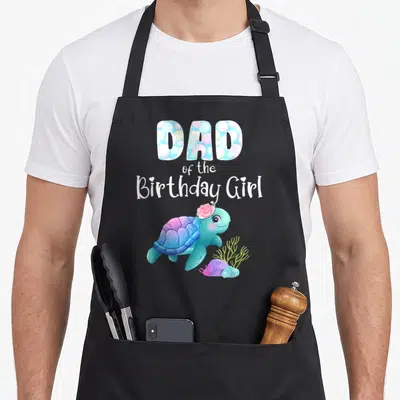 Turtle birthday apron - DAD of the Birthday Girl text with a cartoon sea turtle wearing a pink flower in blue — shown on a black cotton canvas apron against a white background