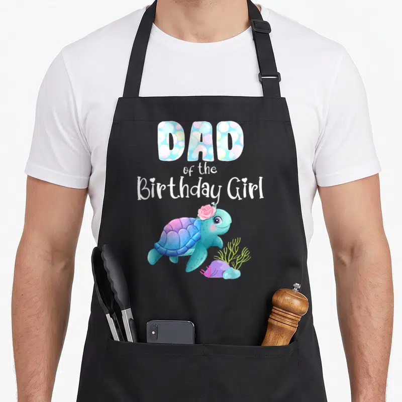 Turtle birthday apron - DAD of the Birthday Girl text with a cartoon sea turtle wearing a pink flower in blue — shown on a black cotton canvas apron against a white background