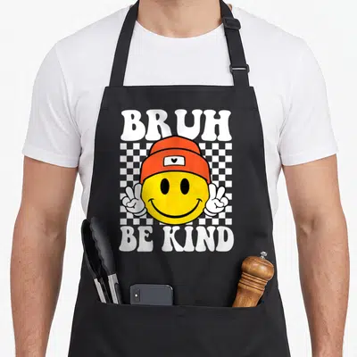 Kind anti unity day apron - White bruh be kind text around yellow smiley in orange beanie making two peace signs — shown on a black cotton canvas apron against a white background
