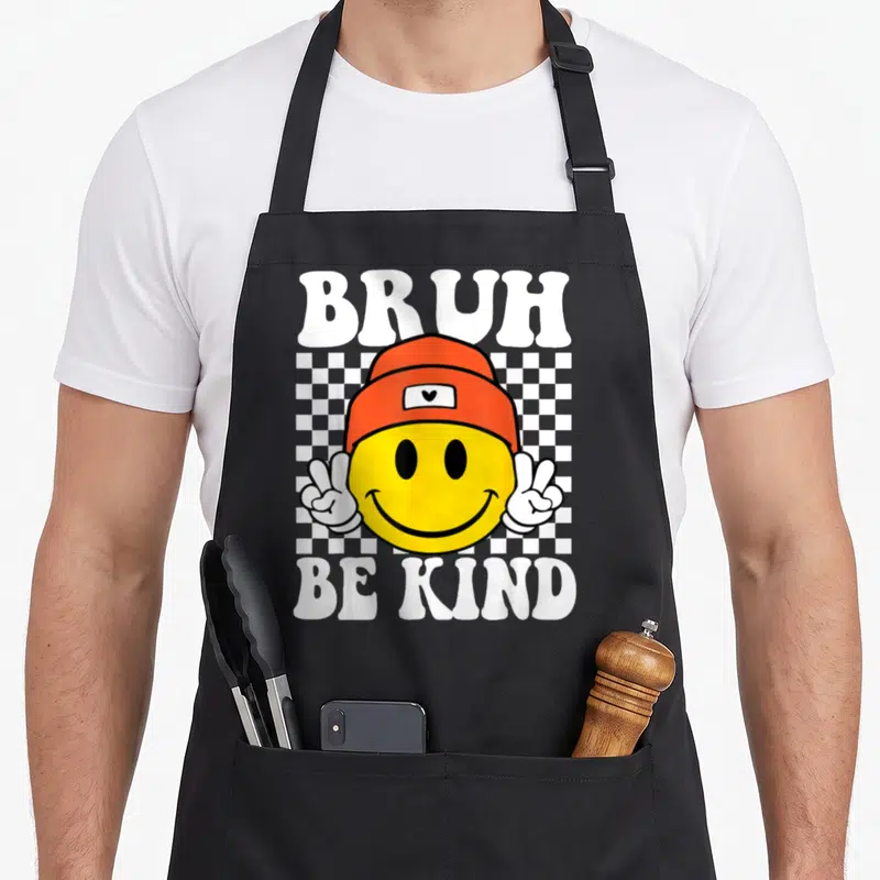Kind anti unity day apron - White bruh be kind text around yellow smiley in orange beanie making two peace signs — shown on a black cotton canvas apron against a white background