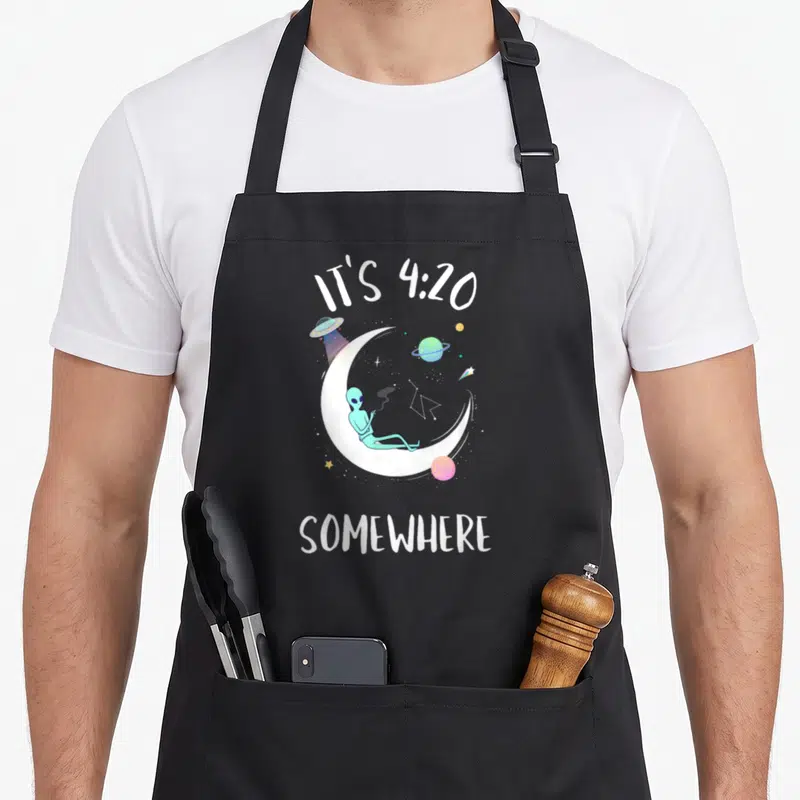 Weed smoking apron - Alien on crescent moon with IT'S 4:20 SOMEWHERE text and space objects — shown on a black cotton canvas apron against a white background