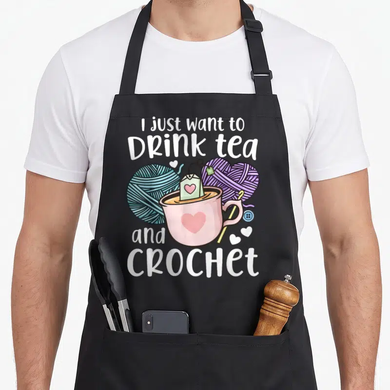 Cute crocheting crochet apron - Pink teacup with blue and purple yarn balls and white text — shown on a black cotton canvas apron against a white background