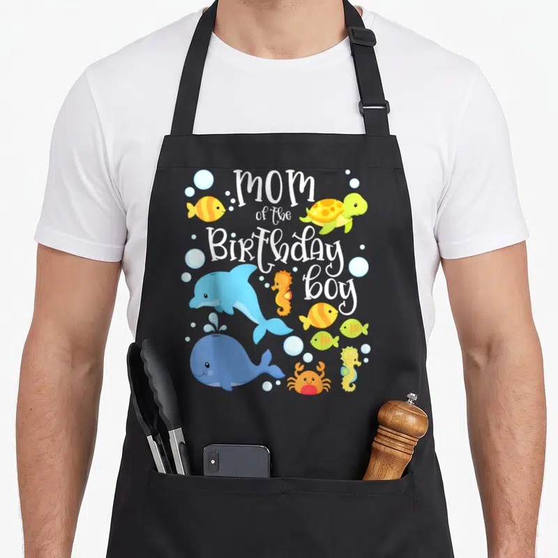 Birthday sea fish apron - Cartoon dolphin whale turtle seahorse crab fish surrounding stacked white text mom — shown on a black cotton canvas apron against a white background