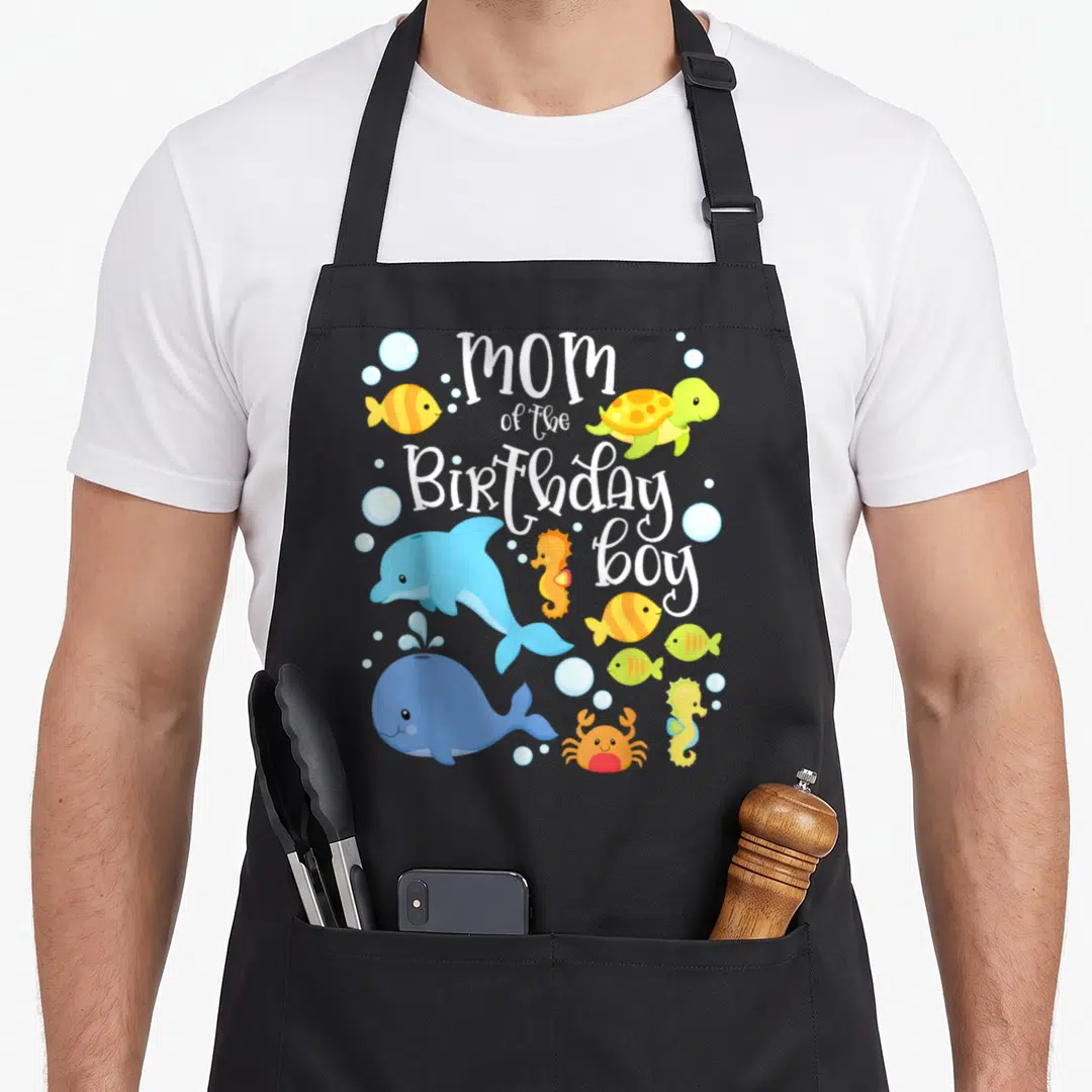 Birthday sea fish apron - Cartoon dolphin whale turtle seahorse crab fish surrounding stacked white text mom — shown on a black cotton canvas apron against a white background