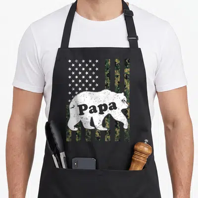 Papa american flag apron - Distressed white bear silhouette containing Papa text over digital camo stripes and white — shown on a black cotton canvas apron against a white background
