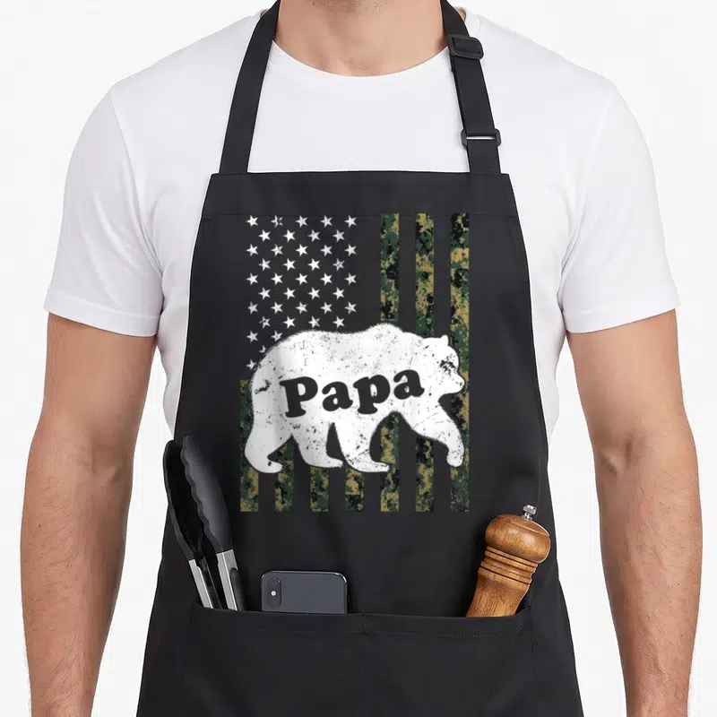Papa american flag apron - Distressed white bear silhouette containing Papa text over digital camo stripes and white — shown on a black cotton canvas apron against a white background
