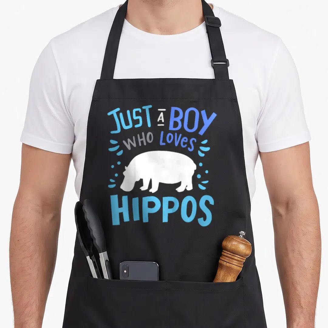Funny hippopotamus hippo apron - White cartoon hippo with blue text Just A Boy Who Loves Hippos and water droplets — shown on a black cotton canvas apron against a white background