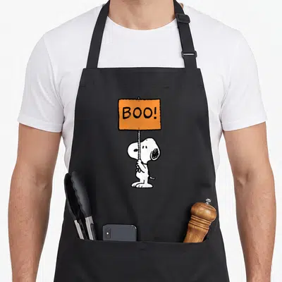 Retro boo halloween apron - Snoopy holding orange sign with BOO! text in white black and orange cartoon style — shown on a black cotton canvas apron against a white background