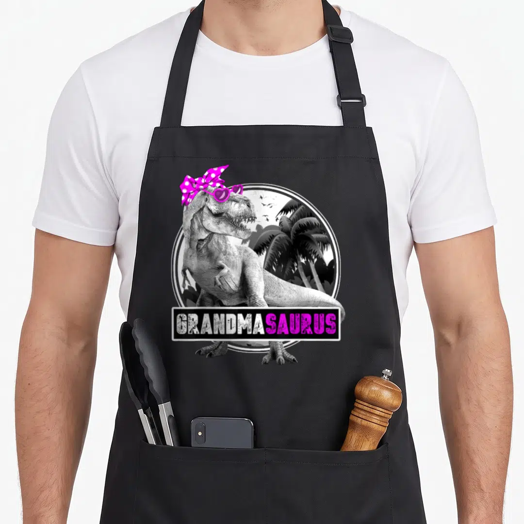 Dinosaur grandma apron - Cartoon T-Rex with pink bow and sunglasses under GRANDMASAURUS text banner — shown on a black cotton canvas apron against a white background