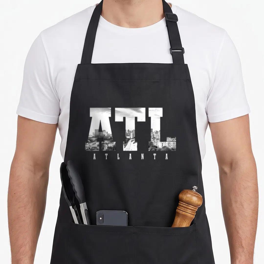 Atl pride apron - ATL letters filled with white and grey cityscape photo above spaced ATLANTA text — shown on a black cotton canvas apron against a white background
