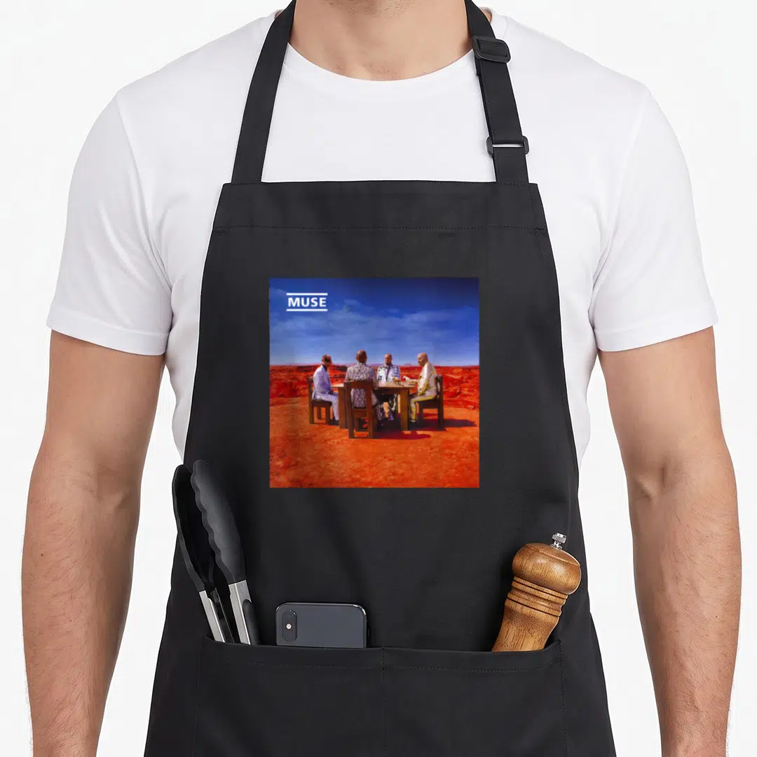 Muse black officially apron - Four men in metallic suits at a wooden table in a red desert with MUSE text in white box — shown on a black cotton canvas apron against a white background