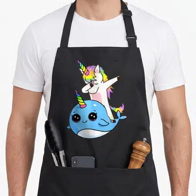 Unicorn friends apron - Cartoon white unicorn dabbing behind a blue narwhal with rainbow horns — shown on a black cotton canvas apron against a white background