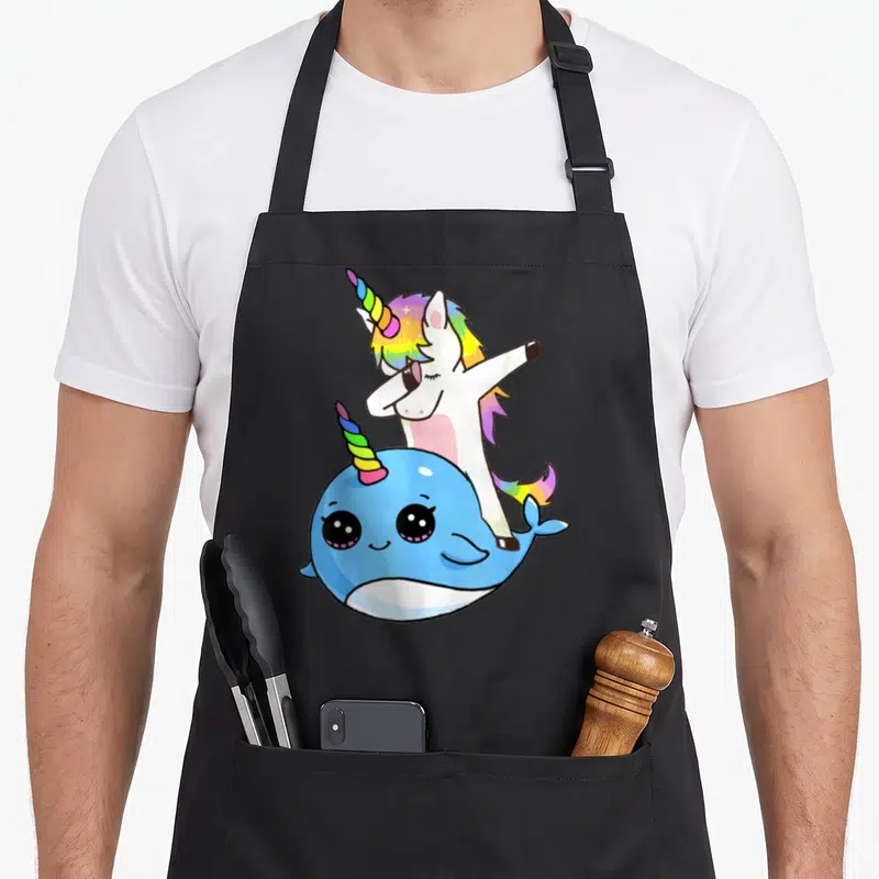 Unicorn friends apron - Cartoon white unicorn dabbing behind a blue narwhal with rainbow horns — shown on a black cotton canvas apron against a white background
