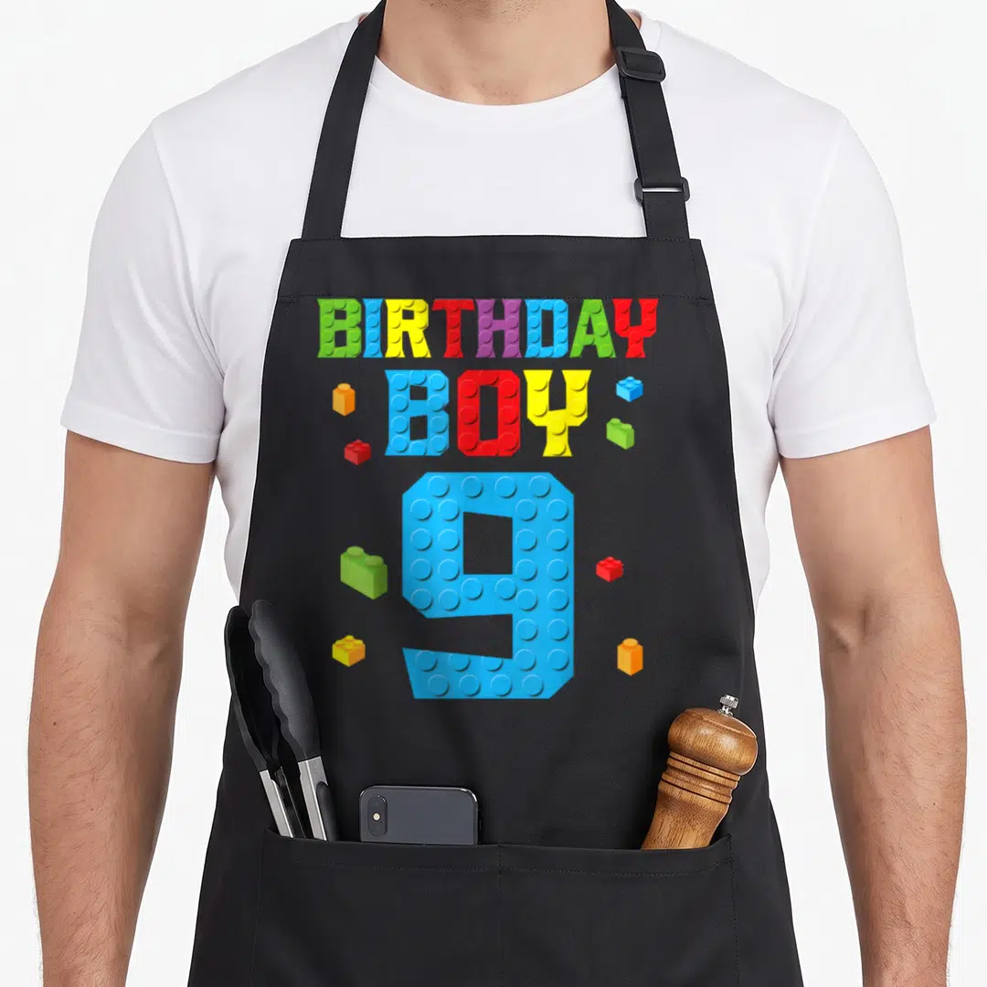 9th building apron - Colorful toy brick text reading BIRTHDAY BOY 9 with scattered blocks — shown on a black cotton canvas apron against a white background
