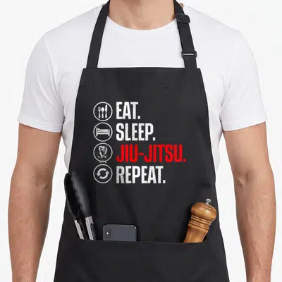 Brazilian jitsu quotes apron - Distressed text Eat Sleep Jiu-Jitsu Repeat with circular icons for food sleep grappling — shown on a black cotton canvas apron against a white background