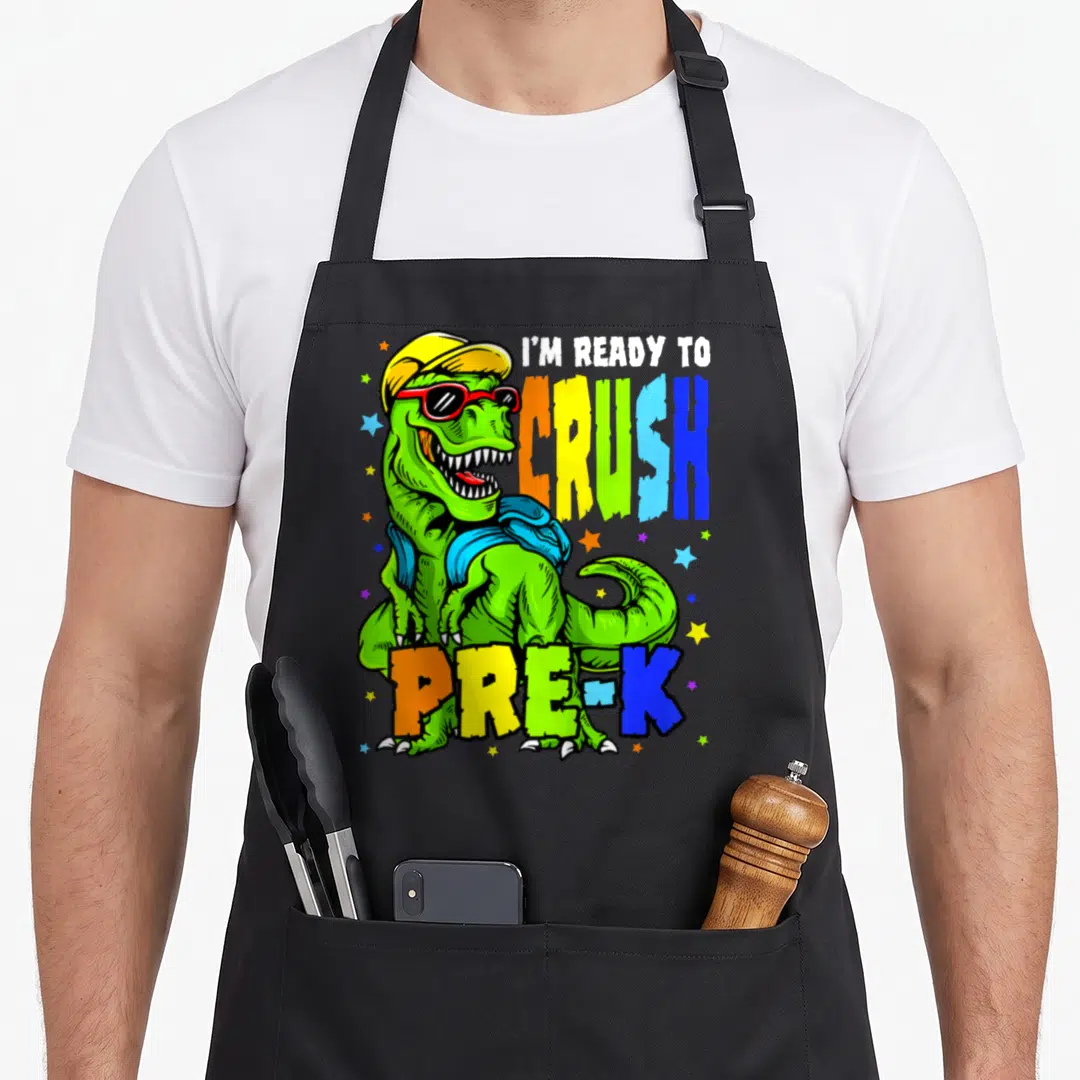 1st dinosaur apron - Cartoon green T-Rex with sunglasses and backpack next to colorful I'M READY TO CRUSH PRE-K text — shown on a black cotton canvas apron against a white background