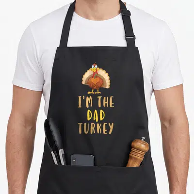 Cool family thanksgiving apron - Cartoon turkey holding pumpkin above I'm The Dad Turkey text — shown on a black cotton canvas apron against a white background